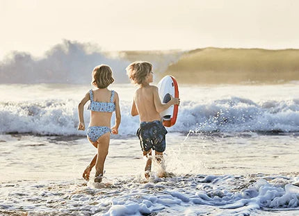 Two children in swimsuits run into the surf on a beach, with one holding a sea scooter