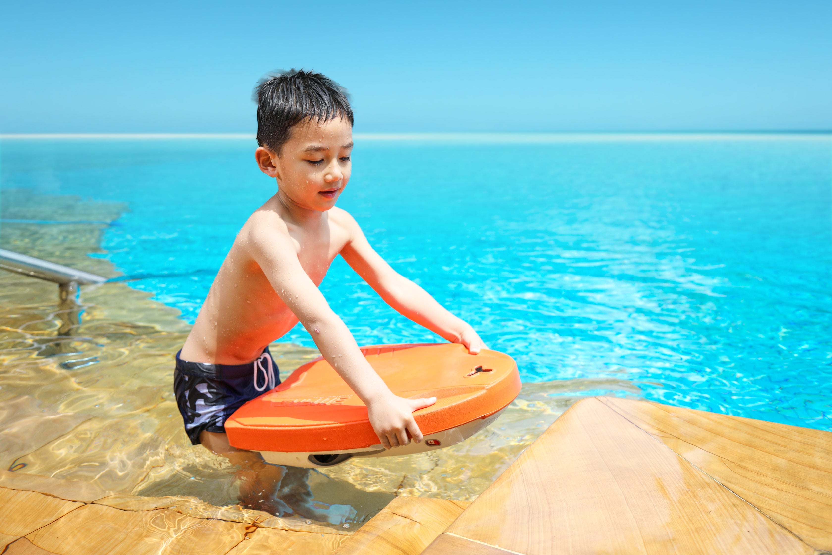 Boy holding orange SUBLUE Swii Electronic Kickboard at poolside, ready for fun.