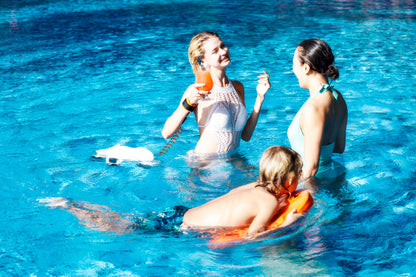 Woman enjoying a drink with a child swimming and another adult in a blue pool.